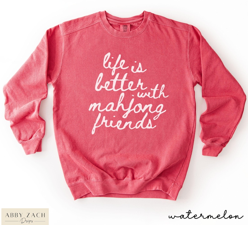 May include: A watermelon-colored sweatshirt with the phrase "life is better with mahjong friends" in white script. The long-sleeved crewneck shirt has a relaxed fit and is made of a soft material. The bottom right corner of the shirt has the word "watermelon".