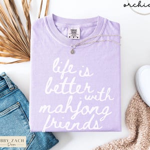 May include: A lavender Comfort Colors t-shirt with the phrase "life is better with mahjong friends" in white script. A silver necklace with a pendant rests on the shirt. The shirt is paired with blue jeans and white sneakers.