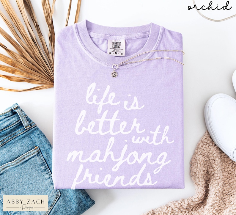 May include: A lavender Comfort Colors t-shirt with the phrase "life is better with mahjong friends" in white script. A silver necklace with a pendant rests on the shirt. The shirt is paired with blue jeans and white sneakers.