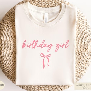 May include: A cream-colored t-shirt with the words "birthday girl" in pink script, accompanied by a pink bow graphic. The shirt is made of a soft material and is perfect for a birthday celebration.