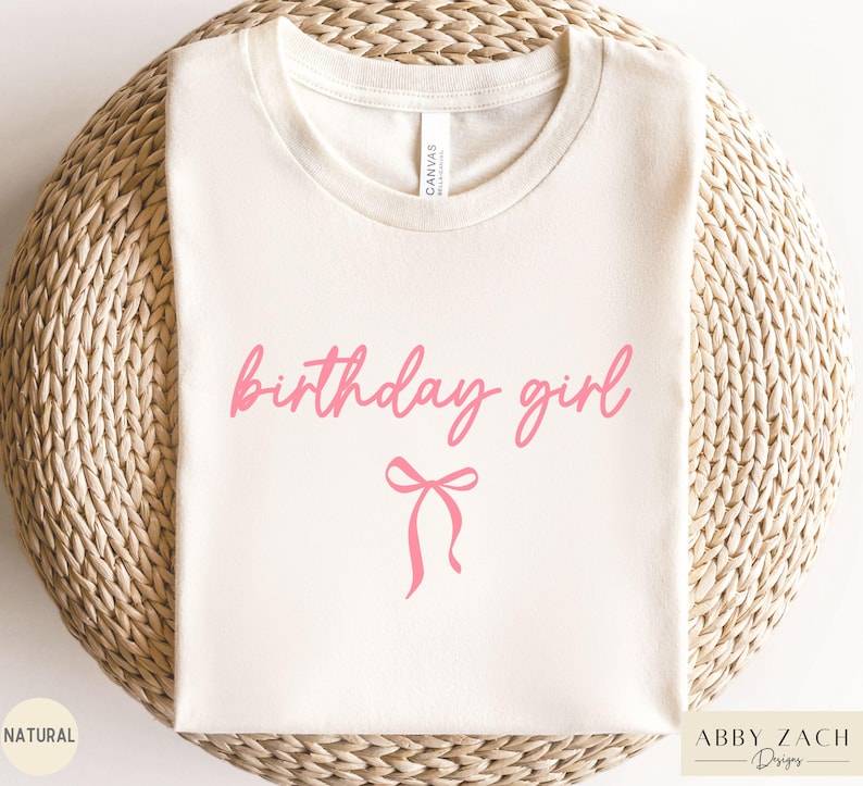 May include: A cream-colored t-shirt with the words "birthday girl" in pink script, accompanied by a pink bow graphic. The shirt is made of a soft material and is perfect for a birthday celebration.