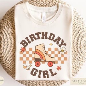 May include: A white t-shirt with a retro-style graphic design featuring a roller skate, dice, and the text "Birthday Girl".