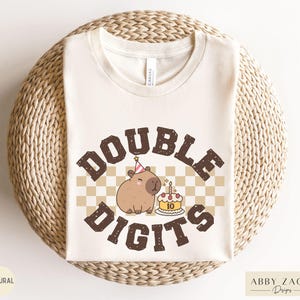 Peut inclure: Un t-shirt blanc avec un capybara brun portant un chapeau de f&ecirc;te et tenant un g&acirc;teau d'anniversaire avec le texte "DOUBLE DIGITS" en lettres brunes sur un fond &agrave; carreaux.