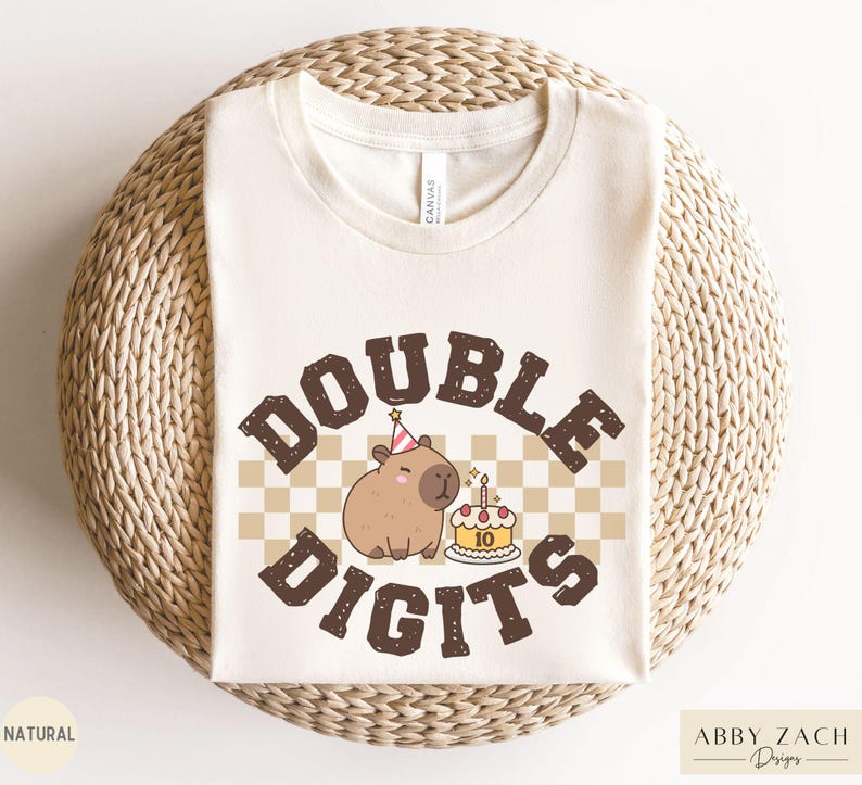Peut inclure: Un t-shirt blanc avec un capybara brun portant un chapeau de f&ecirc;te et tenant un g&acirc;teau d'anniversaire avec le texte "DOUBLE DIGITS" en lettres brunes sur un fond &agrave; carreaux.