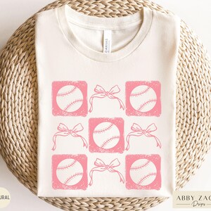 May include: A white t-shirt with a pink design of baseballs and bows. The design is repeated across the shirt.