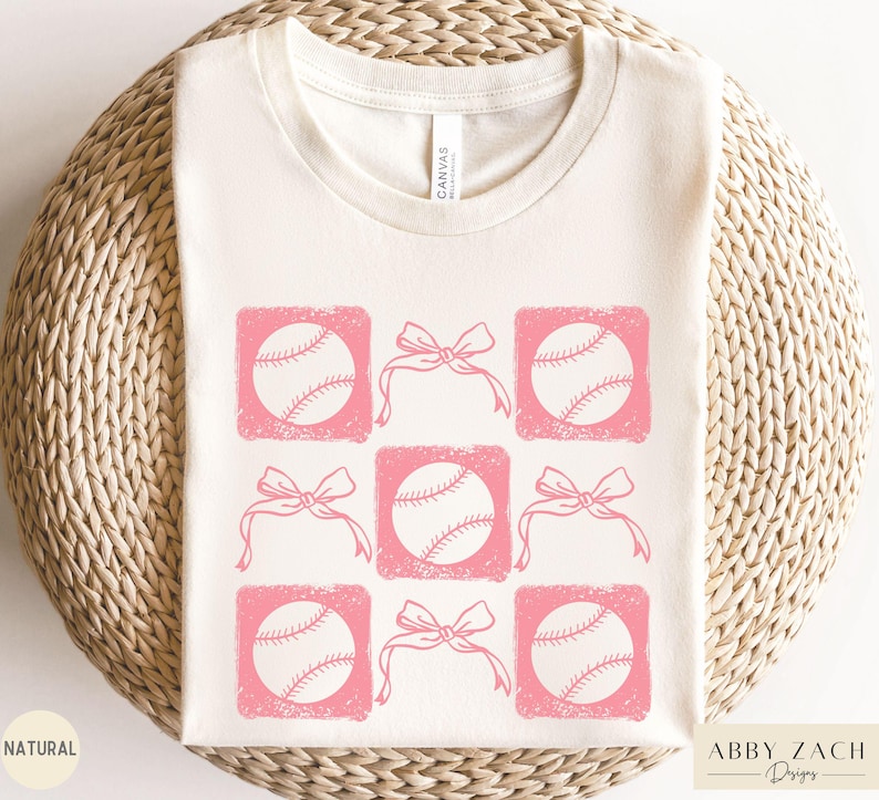 May include: A white t-shirt with a pink design of baseballs and bows. The design is repeated across the shirt.