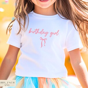 May include: A white t-shirt with the words "birthday girl" in pink script and a pink bow graphic. The shirt is paired with a colorful tutu skirt. The image is of a young person.