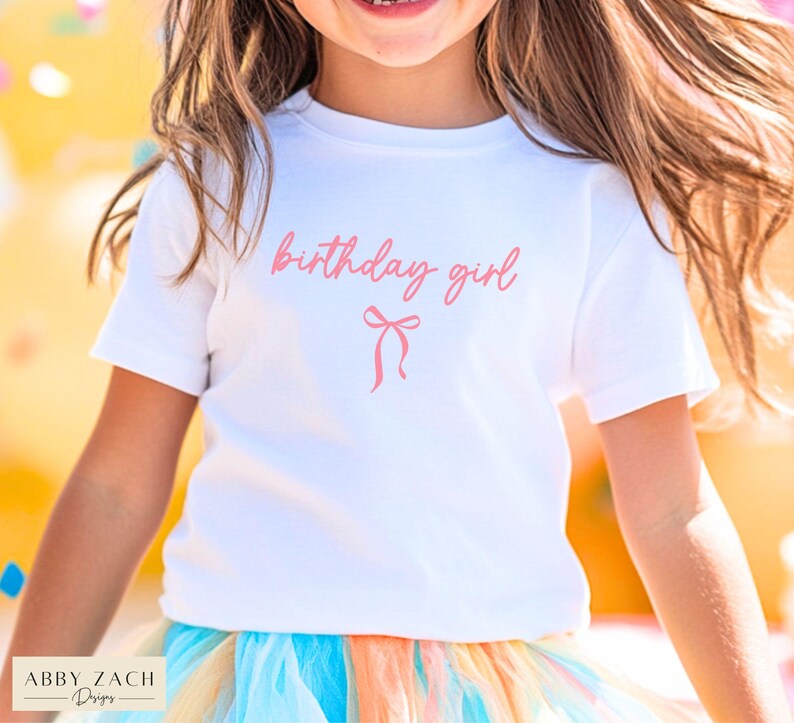 May include: A white t-shirt with the words "birthday girl" in pink script and a pink bow graphic. The shirt is paired with a colorful tutu skirt. The image is of a young person.