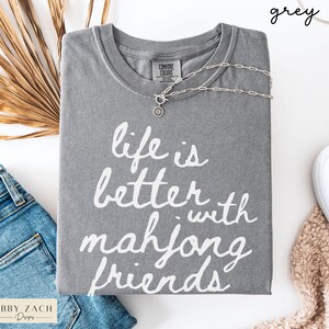 May include: A grey Comfort Colors t-shirt with the phrase "life is better with mahjong friends" in white script. A silver necklace with a circular pendant rests on the shirt. The shirt is paired with blue jeans and a white shoe.