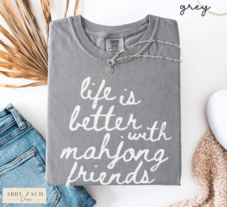 May include: A grey Comfort Colors t-shirt with the phrase "life is better with mahjong friends" in white script. A silver necklace with a circular pendant rests on the shirt. The shirt is paired with blue jeans and a white shoe.