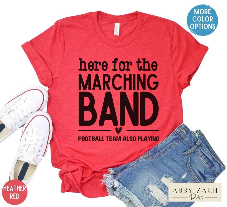 May include: Red t-shirt with the text "here for the marching band" and "football team also playing" in black. The shirt is shown on a model wearing denim shorts.