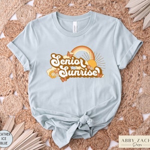 Puede incluir: Una camiseta azul claro con un dise&ntilde;o gr&aacute;fico retro que dice "Senior Sunrise" con un arco&iacute;ris, un sol y mariposas.