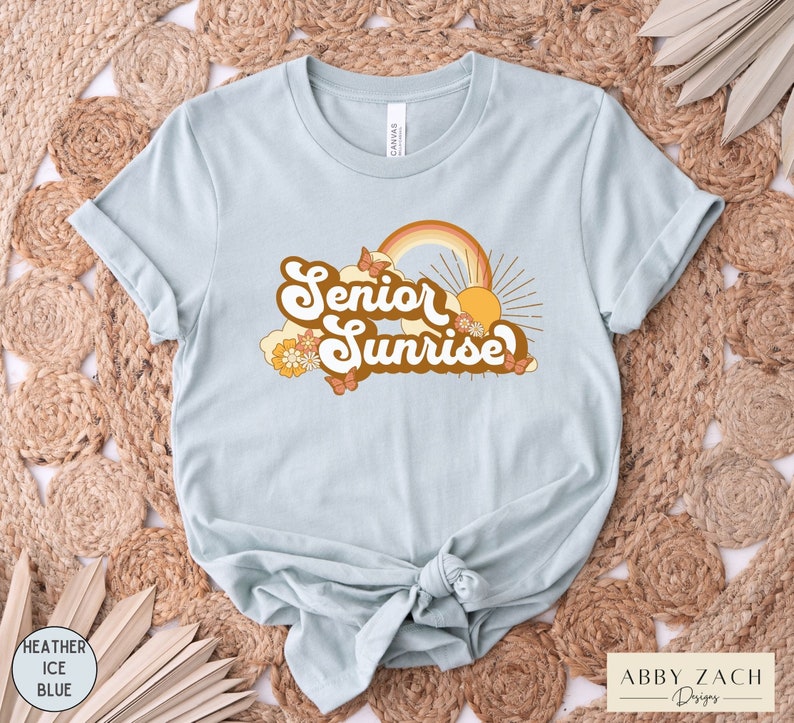Puede incluir: Una camiseta azul claro con un dise&ntilde;o gr&aacute;fico retro que dice "Senior Sunrise" con un arco&iacute;ris, un sol y mariposas.