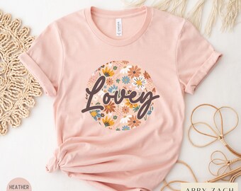 May include: A pink t-shirt with a floral design and the word "Lovey" in a brown script font.
