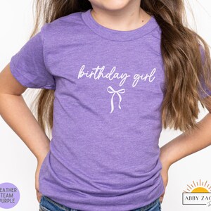 May include: A heather purple t-shirt with the words "birthday girl" in white script and a bow graphic. The shirt is a short-sleeved crew neck. The lower left corner has a purple circle with the words "Heather Team Purple".