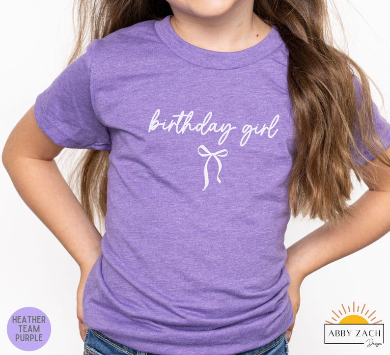 May include: A heather purple t-shirt with the words "birthday girl" in white script and a bow graphic. The shirt is a short-sleeved crew neck. The lower left corner has a purple circle with the words "Heather Team Purple".