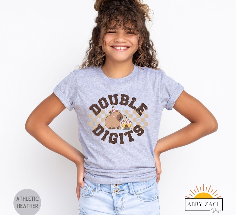 Peut inclure: Un t-shirt gris avec un capybara brun portant un chapeau de f&ecirc;te et tenant un g&acirc;teau d'anniversaire. Le texte "DOUBLE DIGITS" est imprim&eacute; en brun sur le t-shirt.