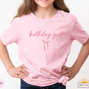 May include: A light pink t-shirt with the words "birthday girl" in a cursive font and a bow graphic. The shirt is made of a soft material and is suitable for a birthday celebration.
