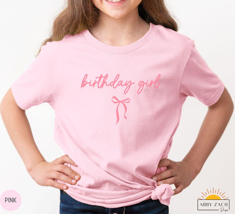May include: A light pink t-shirt with the words "birthday girl" in a cursive font and a bow graphic. The shirt is made of a soft material and is suitable for a birthday celebration.