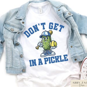 Puede incluir: Camiseta blanca con un pepinillo de dibujos animados azul y verde que lleva una gorra de b&eacute;isbol y sostiene un bate y una pelota de b&eacute;isbol. El texto "Don't Get In A Pickle" est&aacute; impreso en azul en la camiseta.