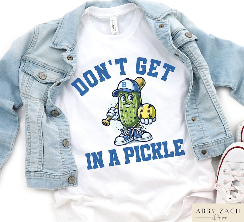 Puede incluir: Camiseta blanca con un pepinillo de dibujos animados azul y verde que lleva una gorra de b&eacute;isbol y sostiene un bate y una pelota de b&eacute;isbol. El texto "Don't Get In A Pickle" est&aacute; impreso en azul en la camiseta.