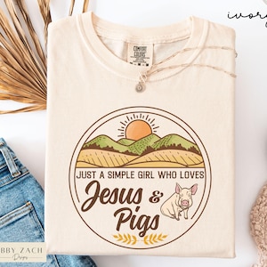 May include: Ivory-colored t-shirt with a round graphic featuring a sun, hills, and a pig. The text reads "JUST A SIMPLE GIRL WHO LOVES Jesus & Pigs." A delicate necklace rests on the shirt. The shirt is made by Comfort Colors.