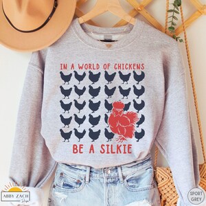 May include: A gray sweatshirt with the text "In a world of chickens be a silkie" printed on it. The text is in black and red, and there are black silhouettes of chickens surrounding the text.