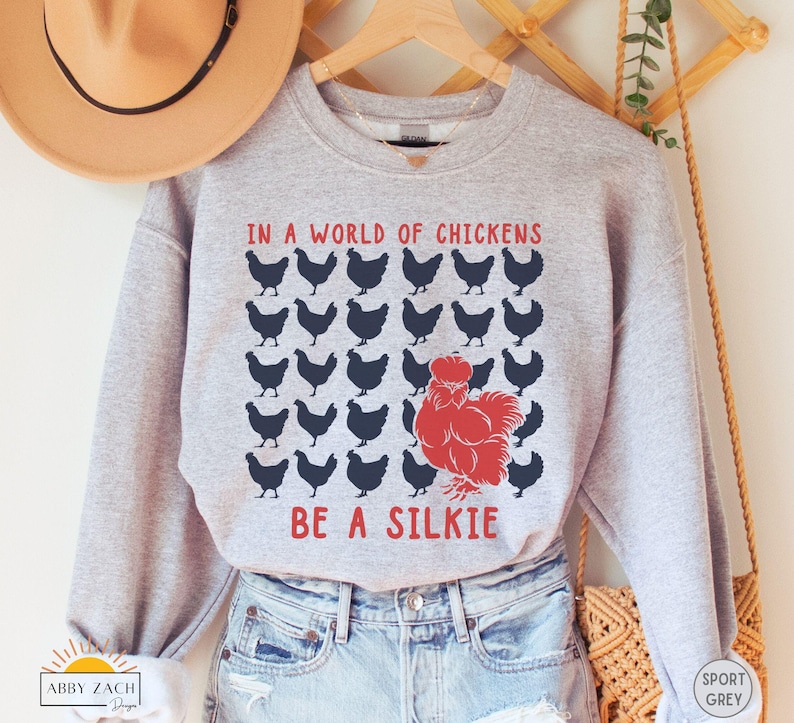 May include: A gray sweatshirt with the text "In a world of chickens be a silkie" printed on it. The text is in black and red, and there are black silhouettes of chickens surrounding the text.
