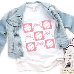 May include: White t-shirt with a pink baseball design. The design features baseballs with pink bows in a grid pattern.