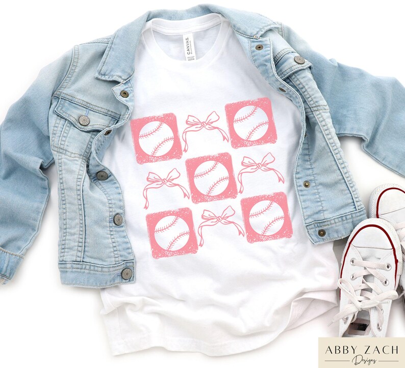 May include: White t-shirt with a pink baseball design. The design features baseballs with pink bows in a grid pattern.