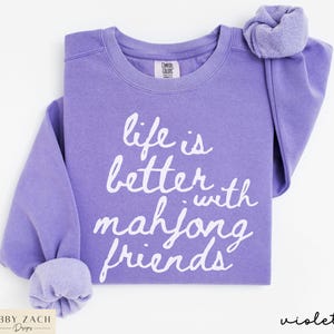 May include: A violet-colored sweatshirt with the text "life is better with mahjong friends" in white script. The sweatshirt has a crew neck and long sleeves with cuffed wrists. The brand label is visible at the top.