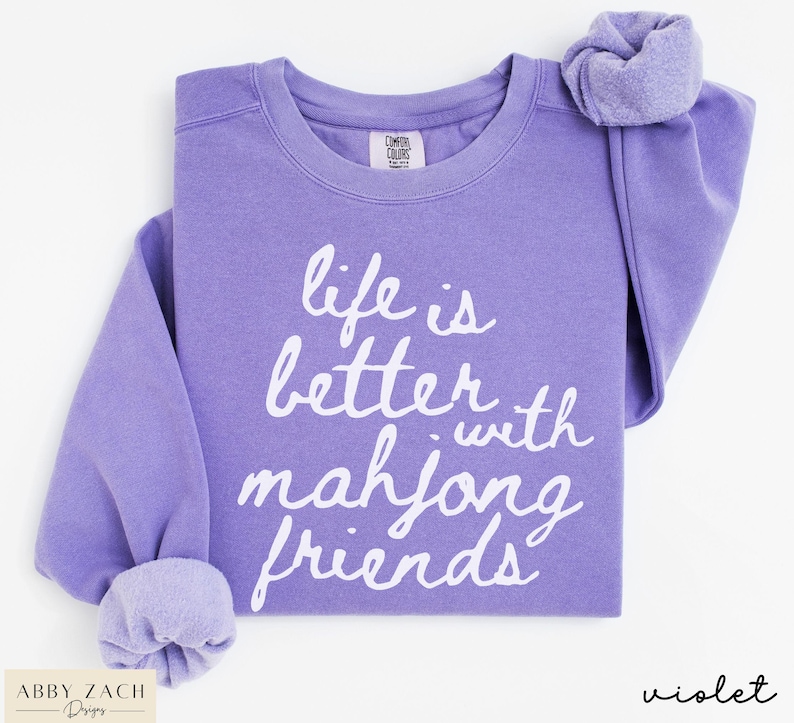 May include: A violet-colored sweatshirt with the text "life is better with mahjong friends" in white script. The sweatshirt has a crew neck and long sleeves with cuffed wrists. The brand label is visible at the top.
