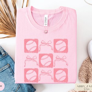 May include: Pink t-shirt with a graphic design of six baseballs with pink bows. The text "Abby Zach Designs" is in the bottom right corner of the image.