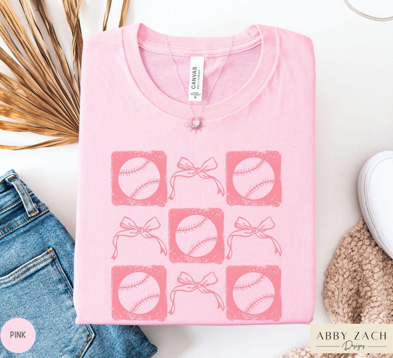 May include: Pink t-shirt with a graphic design of six baseballs with pink bows. The text "Abby Zach Designs" is in the bottom right corner of the image.