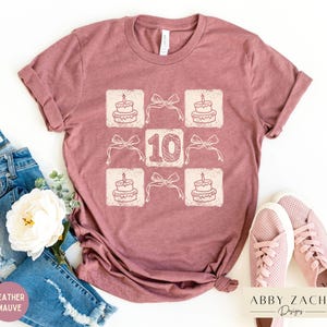 Peut inclure: Un t-shirt en mauve bruy&egrave;re avec un motif graphique repr&eacute;sentant un g&acirc;teau d'anniversaire avec des bougies et le chiffre 10. Le motif est r&eacute;p&eacute;t&eacute; en grille.