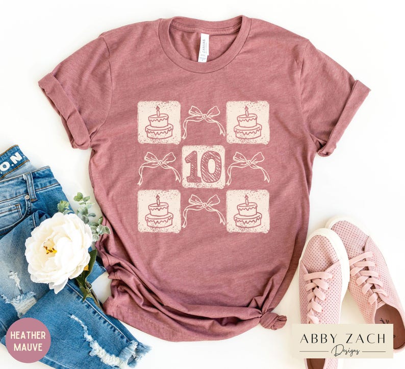 Peut inclure: Un t-shirt en mauve bruy&egrave;re avec un motif graphique repr&eacute;sentant un g&acirc;teau d'anniversaire avec des bougies et le chiffre 10. Le motif est r&eacute;p&eacute;t&eacute; en grille.