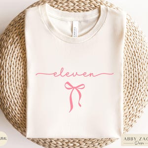 May include: A white t-shirt with the word "eleven" written in cursive script and a pink bow underneath.