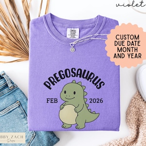 K&ouml;nnte beinhalten: Ein violettes Comfort Colors T-Shirt mit dem Wort "PREGOSAURUS" &uuml;ber einem Cartoon-Dinosaurier. Das Shirt zeigt auch den Monat "FEB" und das Jahr "2026". Eine Halskette und ein Aufkleber "CUSTOM DUE DATE MONTH AND YEAR" sind ebenfalls sichtbar.