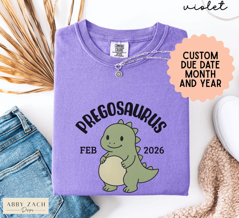 K&ouml;nnte beinhalten: Ein violettes Comfort Colors T-Shirt mit dem Wort "PREGOSAURUS" &uuml;ber einem Cartoon-Dinosaurier. Das Shirt zeigt auch den Monat "FEB" und das Jahr "2026". Eine Halskette und ein Aufkleber "CUSTOM DUE DATE MONTH AND YEAR" sind ebenfalls sichtbar.