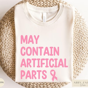 May include: Cream-colored t-shirt with the text "MAY CONTAIN ARTIFICIAL PARTS" in pink, along with a pink ribbon graphic. The shirt is made of a soft material and is folded on a woven surface. The word "NATURAL" is printed on the bottom left.