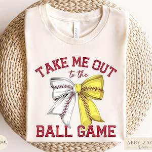 May include: Cream-colored t-shirt with the text "TAKE ME OUT to the BALL GAME" in red. The design features a bow made of a baseball and a yellow ribbon. The shirt is made of a natural fabric.