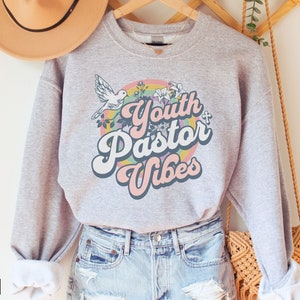 May include: Gray sweatshirt with a rainbow design and the text "Youth Pastor Vibes". The design includes a white dove, flowers, and a cross.