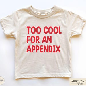 May include: A cream-colored t-shirt with the red text "TOO COOL FOR AN APPENDIX." The shirt has short sleeves and a crew neck. The bottom right corner of the shirt has the text "ABBY ZACH Designs."