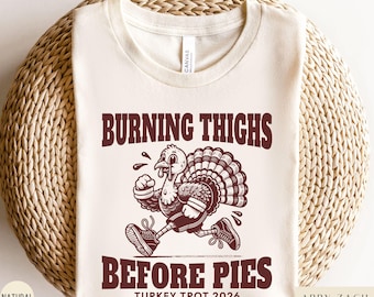 May include: A cream-coloured t-shirt featuring a dark brown graphic of a turkey wearing trainers and running. The text reads BURNING THIGHS BEFORE PIES and TURKEY TROT 2026 in a bold, dark brown font. The shirt has a crew neckline and short sleeves.