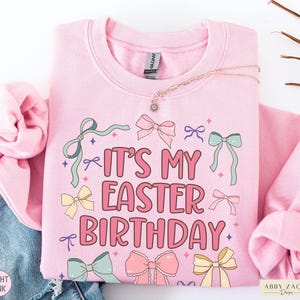 May include: Light pink sweatshirt with the text "IT'S MY EASTER BIRTHDAY" in pink and white. The design includes pastel bows and stars. A delicate necklace with a sun pendant rests on the sweatshirt.