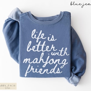 May include: A blue jean colored sweatshirt with the text "life is better with mahjong friends" in white script. The sweatshirt has long sleeves and a crew neck. The brand "Abby Zach Designs" is in the bottom left corner.