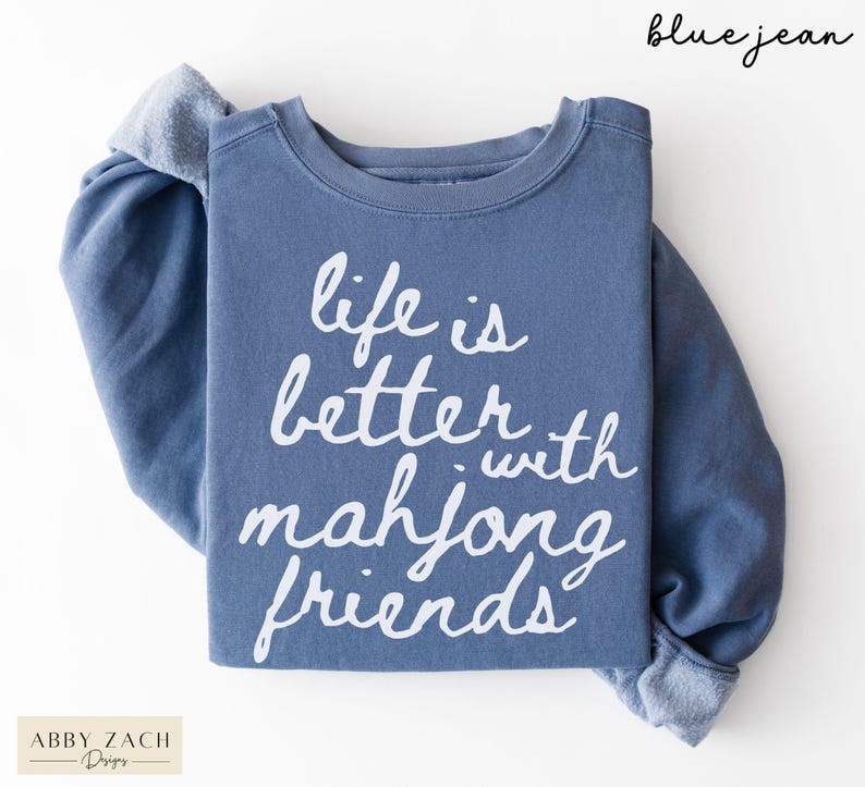 May include: A blue jean colored sweatshirt with the text "life is better with mahjong friends" in white script. The sweatshirt has long sleeves and a crew neck. The brand "Abby Zach Designs" is in the bottom left corner.