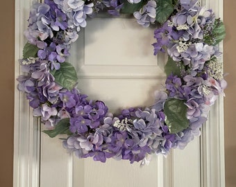 May include: A purple and white hydrangea wreath with green leaves. The wreath is hanging on a white door.