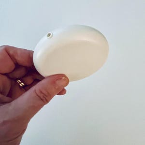 May include: A smooth, white, oval-shaped object with a small hole at the top. The object is held in a hand with a gold ring. The background is a plain white wall. The object appears to be a decorative item or a small container.