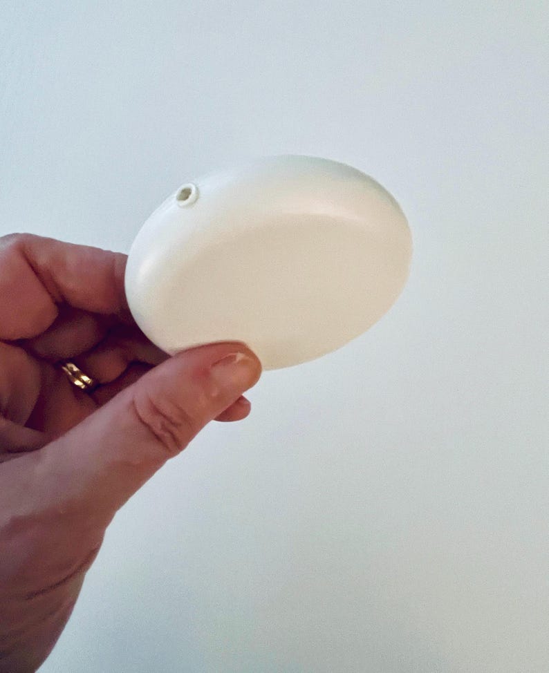 May include: A smooth, white, oval-shaped object with a small hole at the top. The object is held in a hand with a gold ring. The background is a plain white wall. The object appears to be a decorative item or a small container.